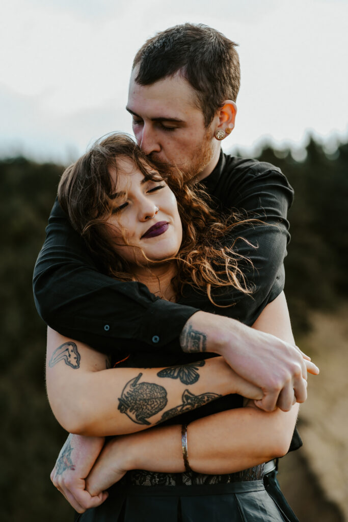  A couple in all black wedding attire, covered in beautiful tattoos embrace, facing their photographer. The groom wraps his arm around the bride from behind as the wind blows through her hair. 