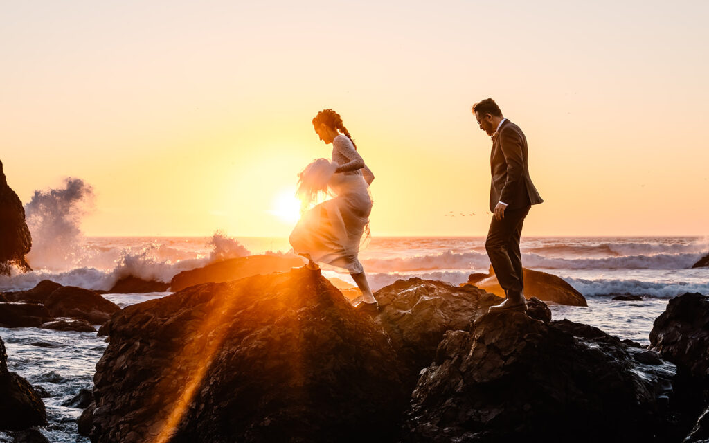 After considering elopement vs wedding, a couple in wedding attire climb a sea-rock. The ocean waves roll behind them. The sun is setting behind the ocean and casts deep orange and soft pink rays of light