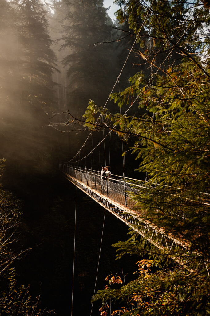 After considering elopement vs wedding, a couple embraces in the middle of a suspended bridge that is surrounded by greenery from the forest. there is a light haze in the top left corner and catches the golden color of the sunrise. 