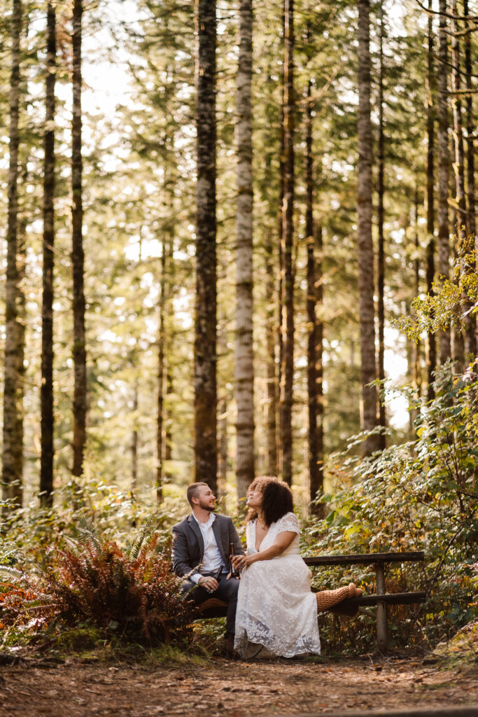 After considering elopement vs wedding, a couple in wedding attire shares a beer at a picnic table in the woods as morning light leaks through the foliage of a deep coastal forest canopy.