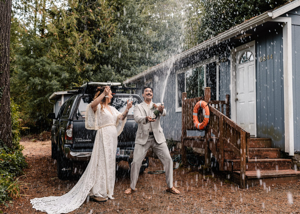 A couple in wedding attire sprays freshly popped champagne in celebration as they prepare to head to their elopement destination 
