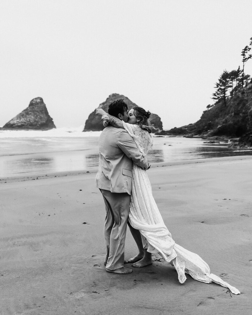 After planning their elopement timeline, a couple in wedding attire embraces and kiss on the beach after their ceremony. Wind blows through their hair and makes their wedding attire billow as they kiss on the rocky Oregon Coast 