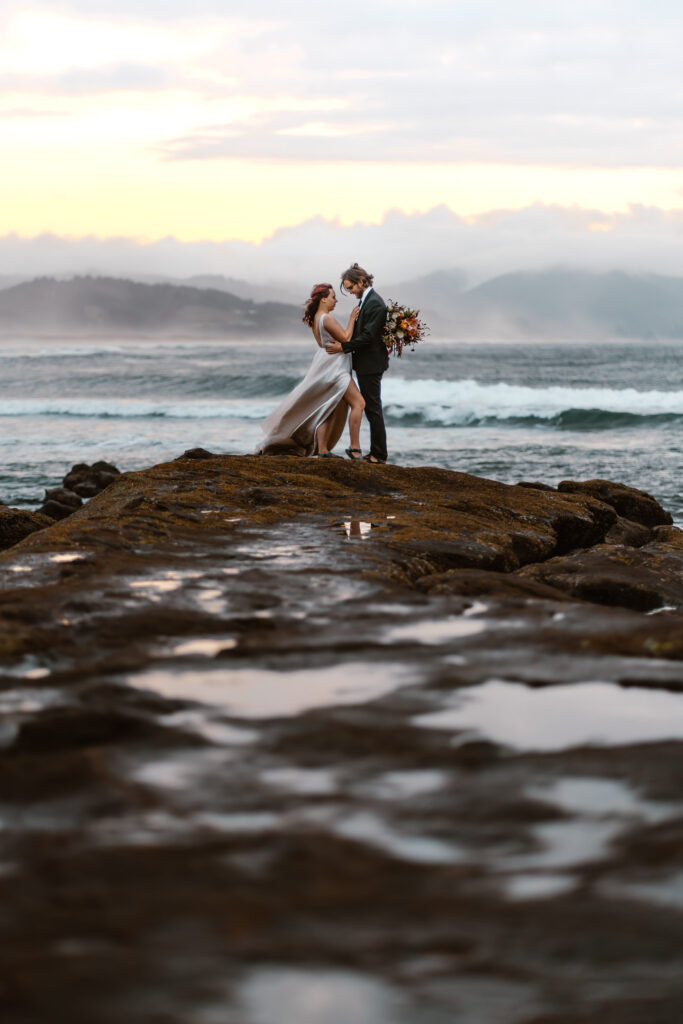 After choosing to get married without a wedding, a bride and groom embrace on a seaside cliff. Blue waves from the pacific roll behind them, clouds, low fog,  and rolling green hills can be seen in the distance as they embrace.