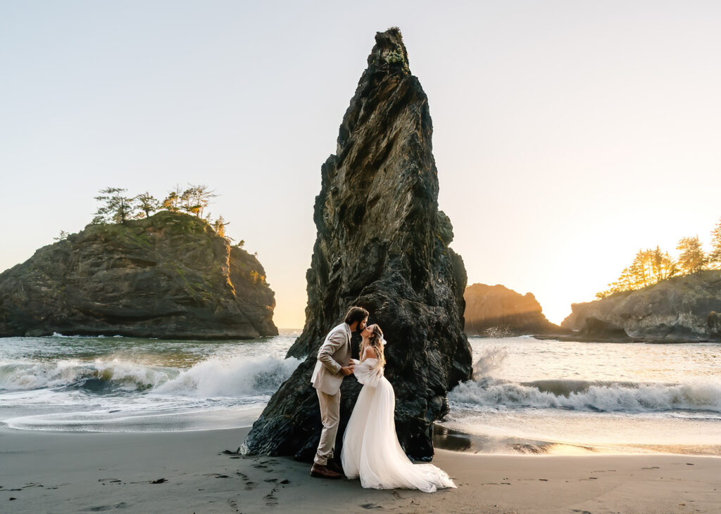 During their elopement timeline, a couple in wedding attire kiss in front of a jagged sea rock as waves crash behind them and the sunset casts a golden light on the rocky coast.