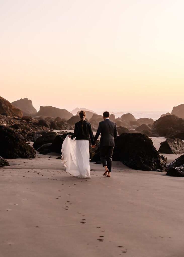 After planning their elopement timeline, a couple walks along the rocky beach as the sunsets in the distance. The are surrounded by soft pink and gold light and wet footprints in the sand show the path they have taken. 