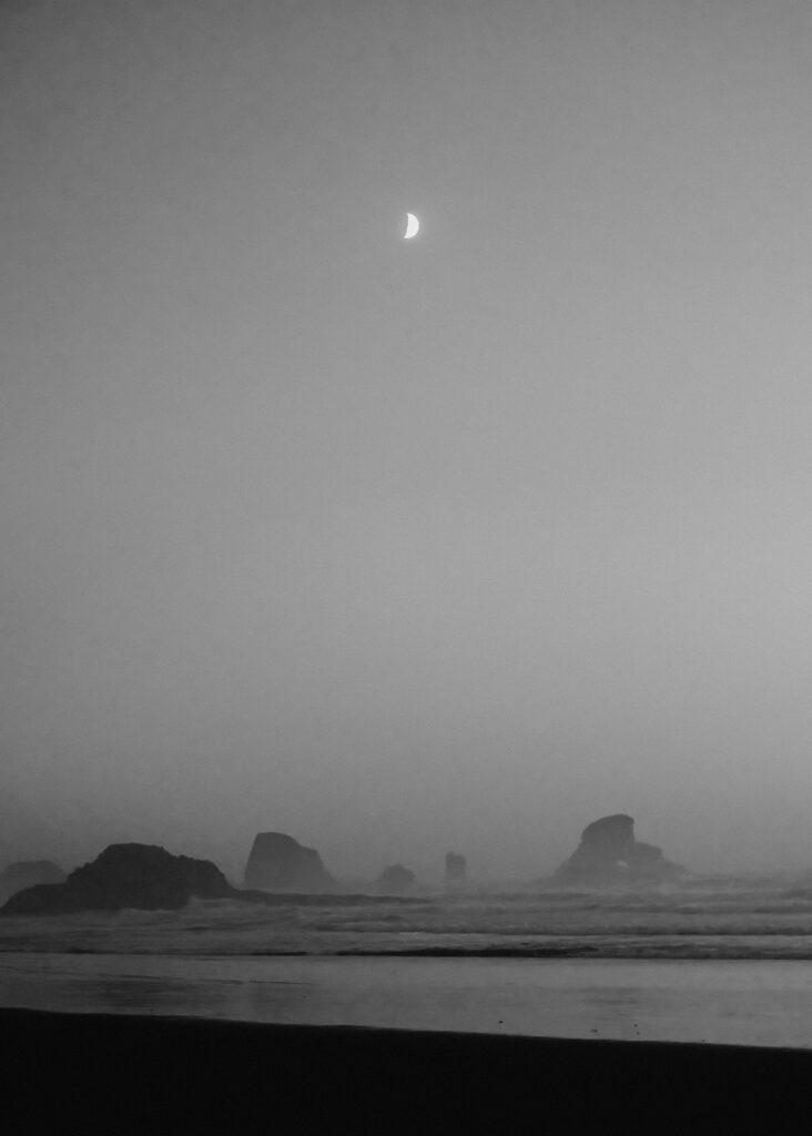 A bright, crescent moon is perfectly centered over a rocky coastline. A light fog creeps in behind the tide.