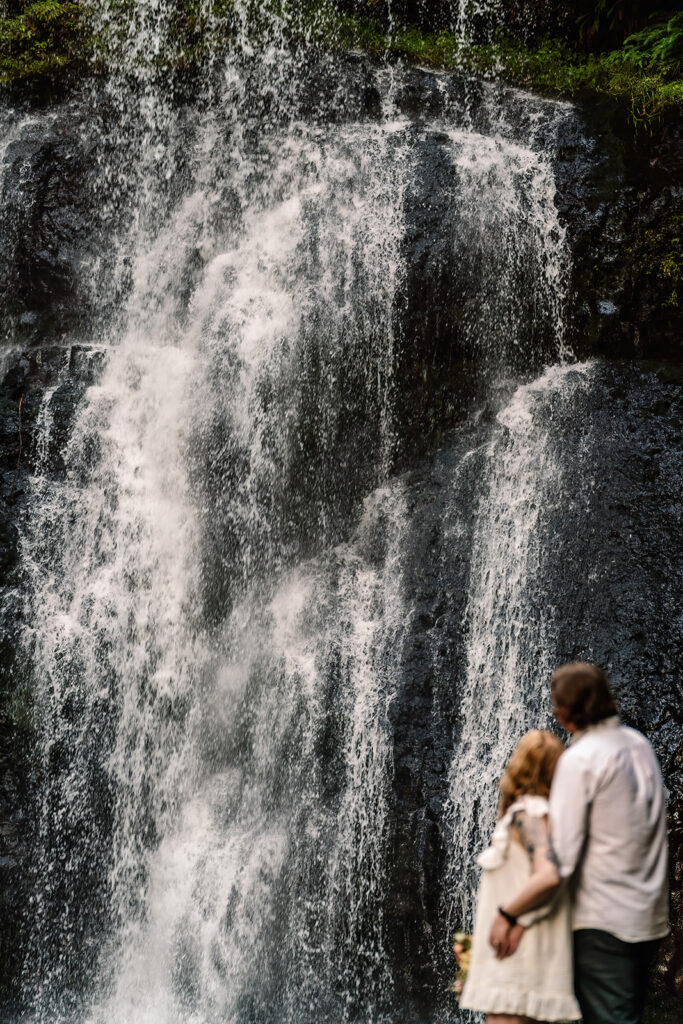 To begin their elopement timeline, a couple embraces and look out towards a cascading waterfall
