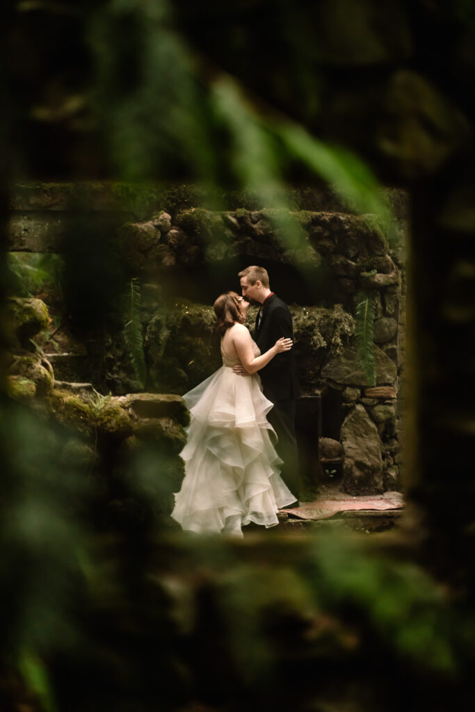 After choosing to get married without a wedding, a bride and groom share their first kiss as a married couple in the remnants of an old stone house, covered in moss. This moment is framed up and captured through the frame of a window overgrown with bright green ferns.