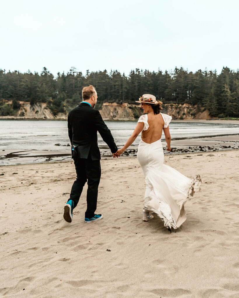 A couple walks hand in hand after choosing to get married without a wedding. They look at each other as they walk along a sandy beach surrounded by cliffs and trees. The wind makes her elopement dress billow as they walk.