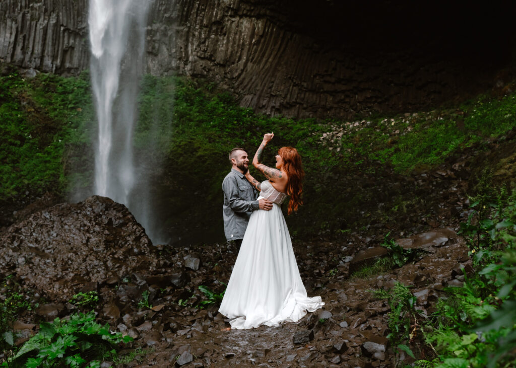After choosing to get married without a wedding, a couple cheers in their wedding attire. They are surrounded by lush greenery, basalt rock, and a gushing waterfall behind them. 