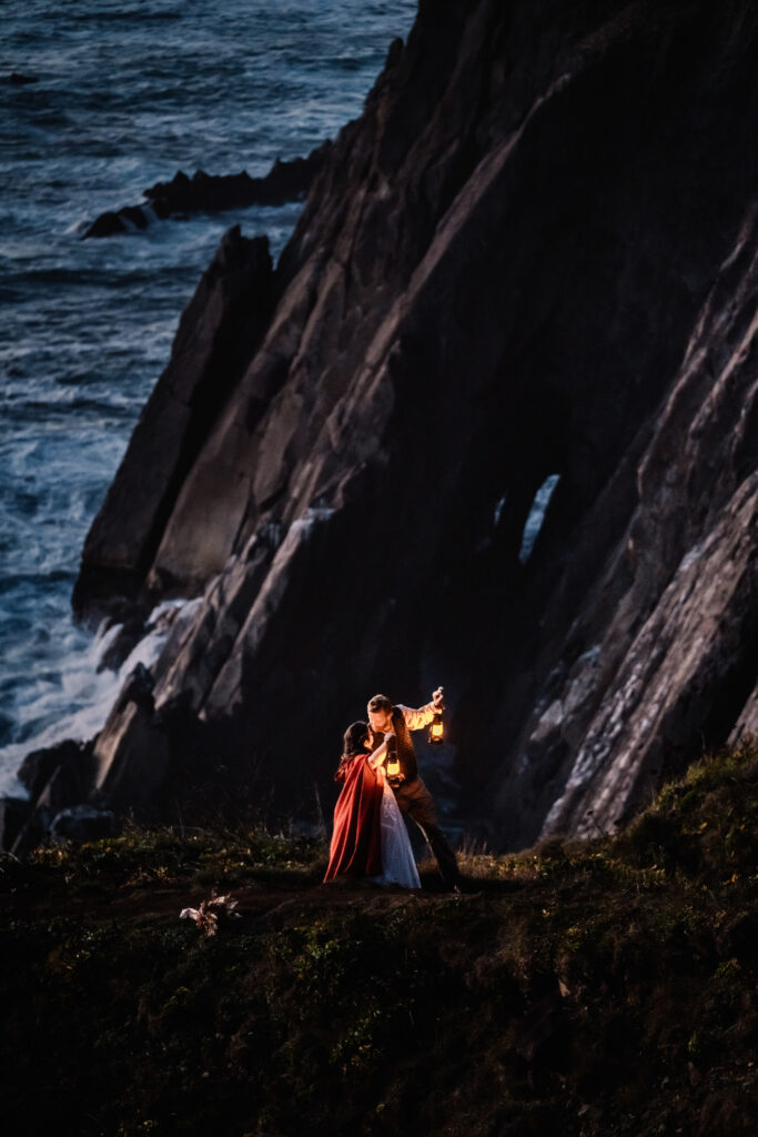 After choosing to get married without a wedding, a couple in wedding attire holds lanterns on the cliffside as the sun sets. Waves can be seen crashing below them and jagged rocks of basalt form their background.