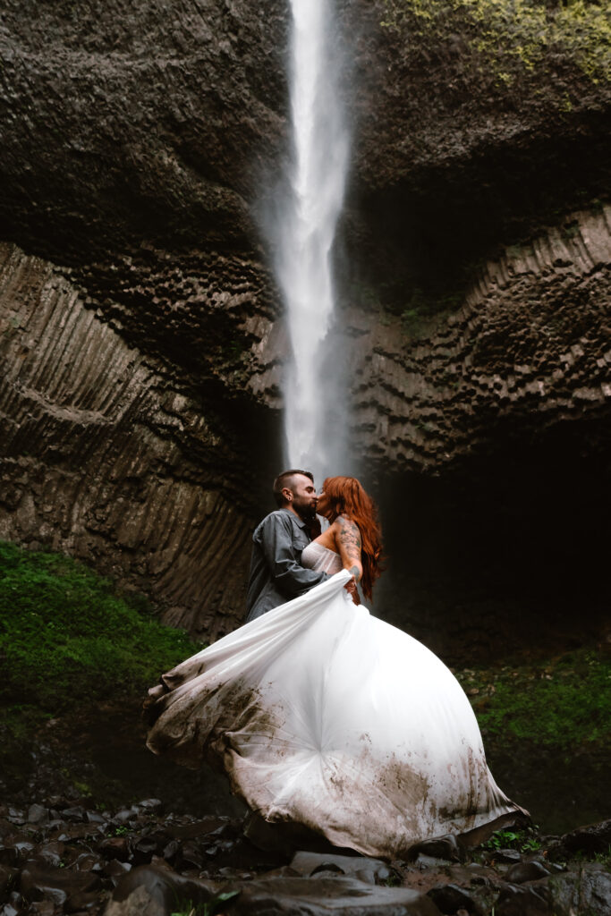 After choosing to get married without a wedding, a couple kisses in front of a gushing waterfall. The brides white dress billows around her and the bottom of it is painted with dark mud.