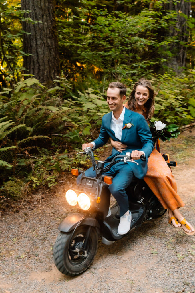 After choosing to get married without a wedding, a couple in vintage attire ride through a lush, green forest on a vintage scooter. The bride sits side-saddle behind her groom, holding her bouquet. They both grin as they zoom by. 