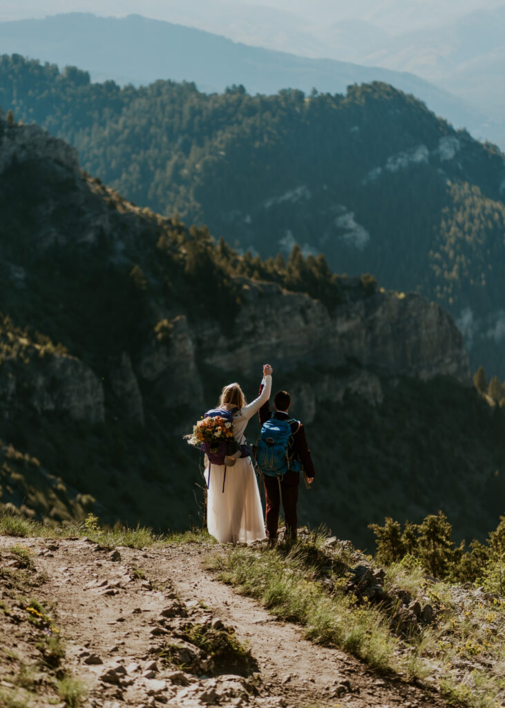 After choosing to get married without a wedding, a couple in wedding attire cheer as they look out to the ridge line as they hike down a switchback. They wear backpacking bags over their attire and the bride keeps her bright bouquet in her backpack. 