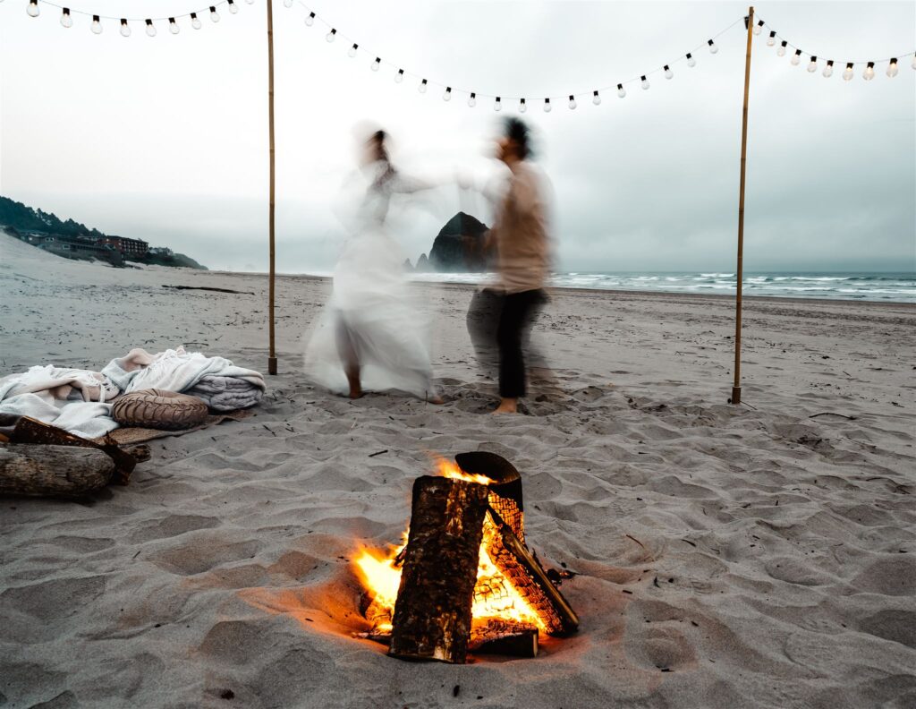 After choosing to get married without a wedding, A couple celebrates in their wedding attire. They are blurry as they dance in front of a glowing campfire. Cozy pillows and blankets surround them, and the iconic Haystack rock of Cannon Beach is framed in the background between them. 