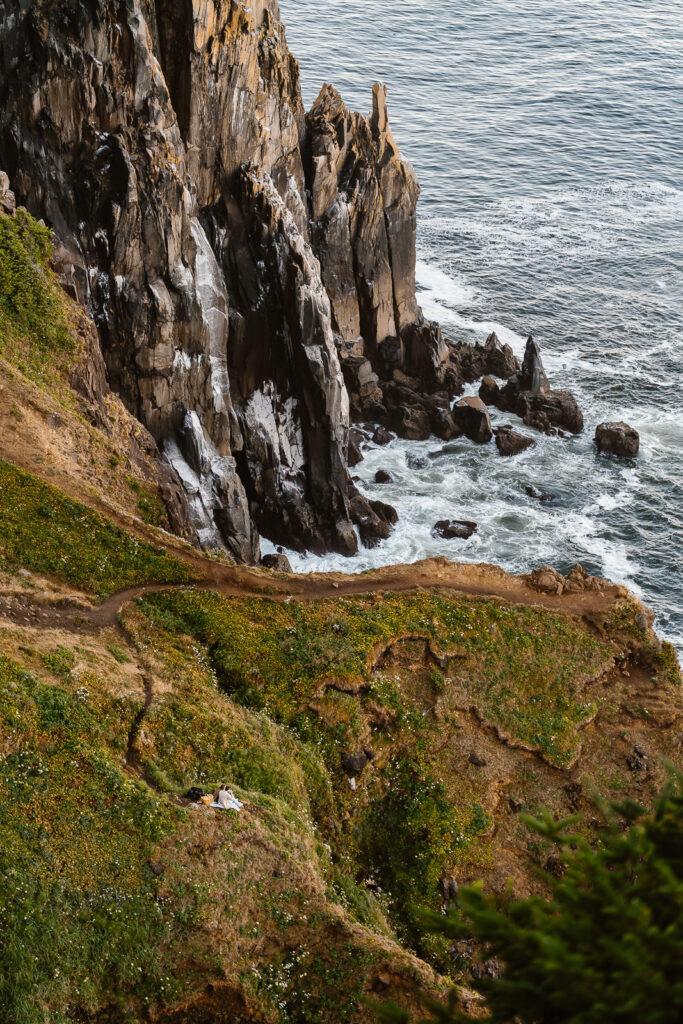 A landscape shot of where this couple began their morning adventures for their coastal elopement. they snuggle as they gaze over a coastal cliff into the deep blue ocean. They are tiny in the landscape. 
