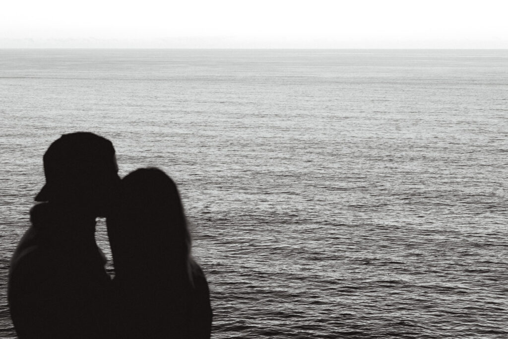 a couple embraces with gentle waves behind them during the morning of their coastal elopement. the focus of the image is the texture of the sea as their blurred bodies frame it.