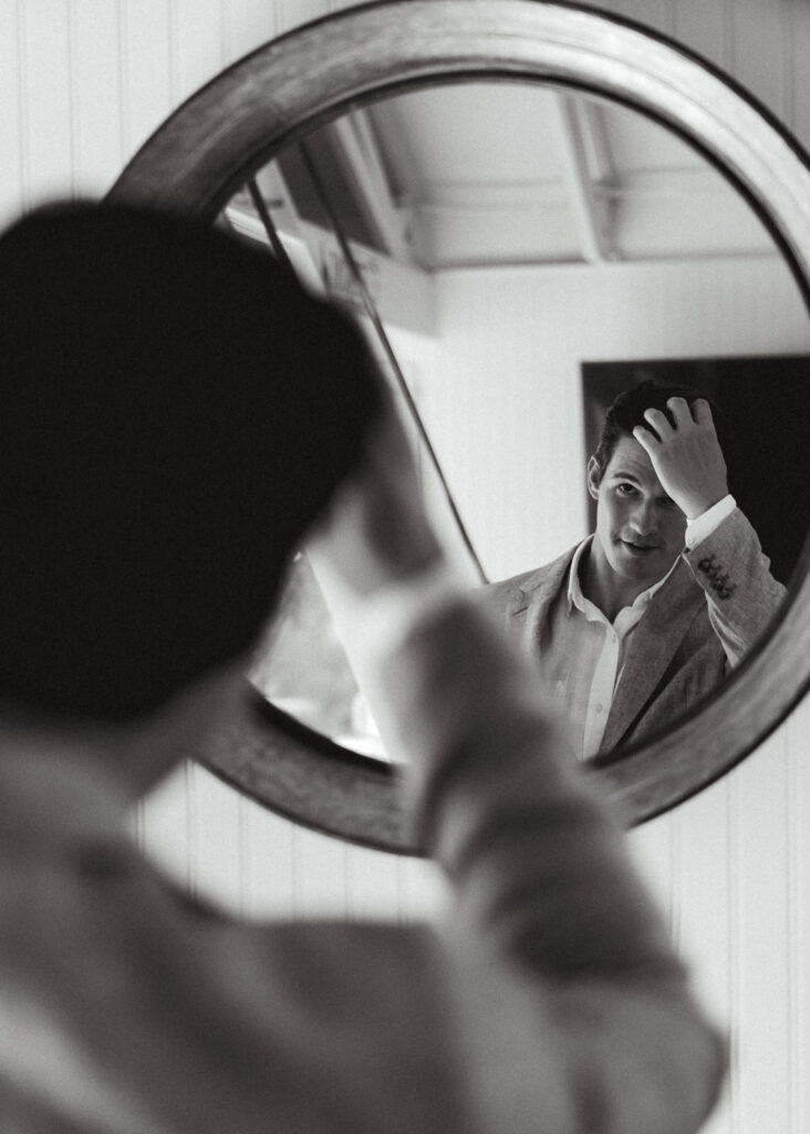 A black and white image of a groom getting ready for his coastal elopement. He gazes in the mirror 