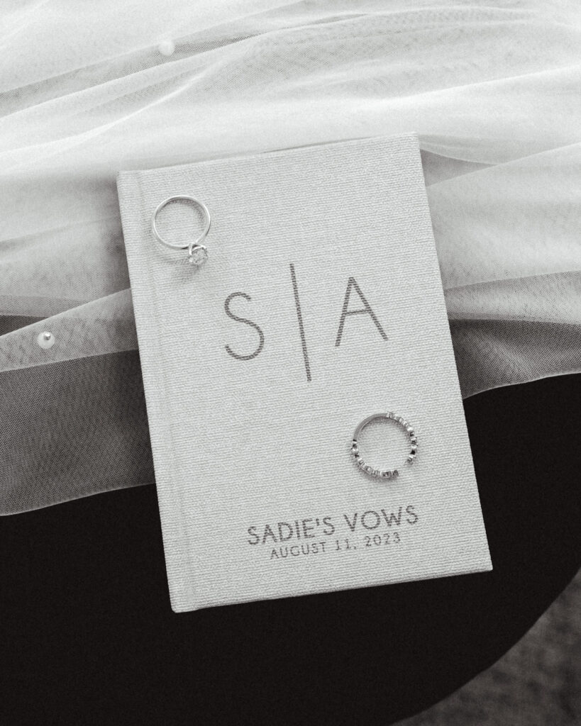 A black and white image of a brides details for her coastal elopement. A vow book lays on top of her pearled veil, her rings are placed on top of it. 