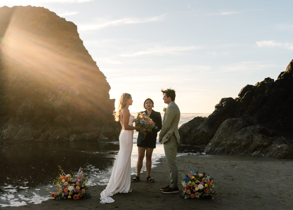 A couple exchanges vows during their coastal elopement as the sun flares behind them