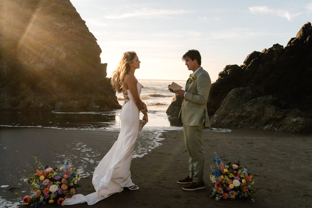 During their coastal elopement, a bride and groom exchange hand written vows in front of rocky features on the beach during sunset.
 