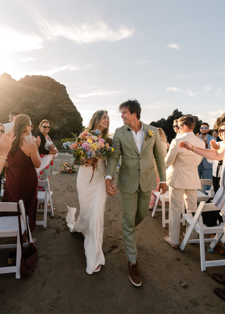a bride and groom exit their ceremony of their coastal elopement on a rocky beach during sunset. Their friends and family cheer behind them. 