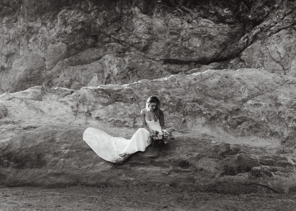 during her coastal elopement, a bride sits on a large sea rock gazing down at her bouquet.