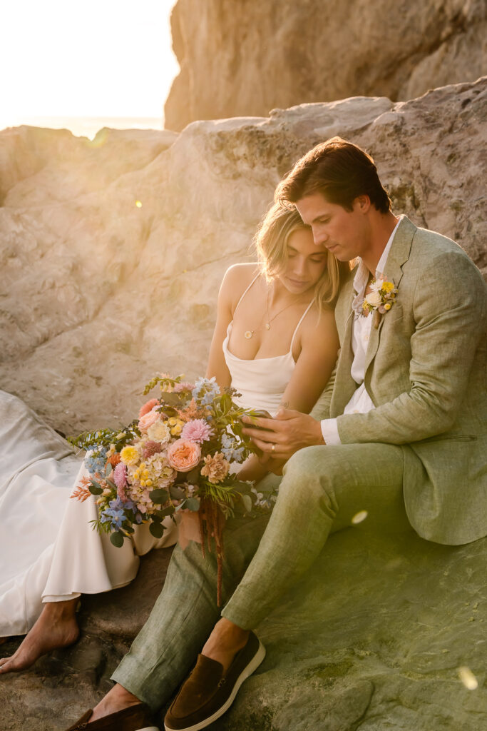 A bride and groom sit next to each other during their coastal elopement. They embrace as the golden sun dips behind them.