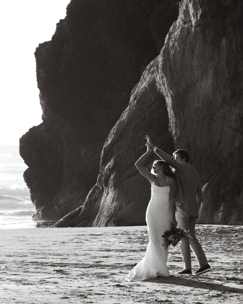 A black and white photo of a bride and groom dancing on a rugged beach during their coastal elopement.