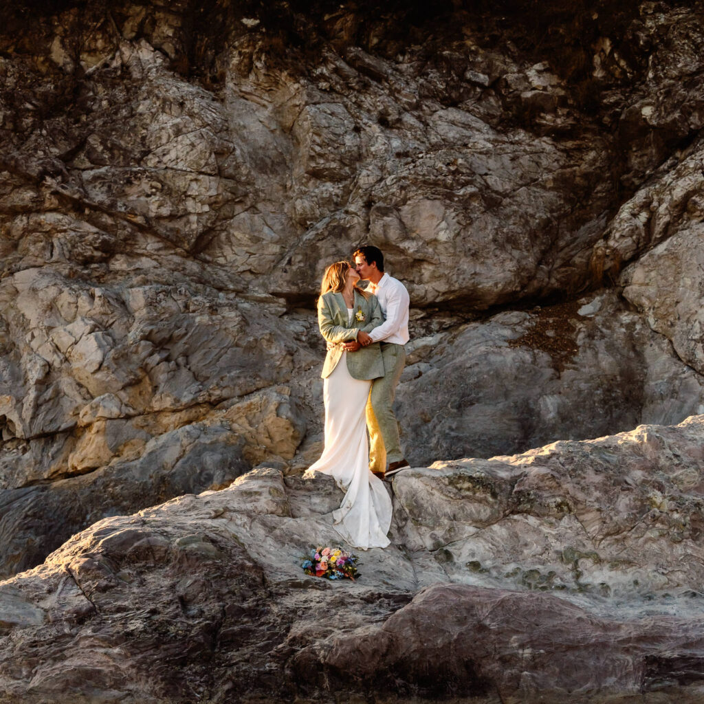 a bride and groom embrace on a large sea rock during their coastal elopement. She wears his coat and her bouquet lays beneath them.