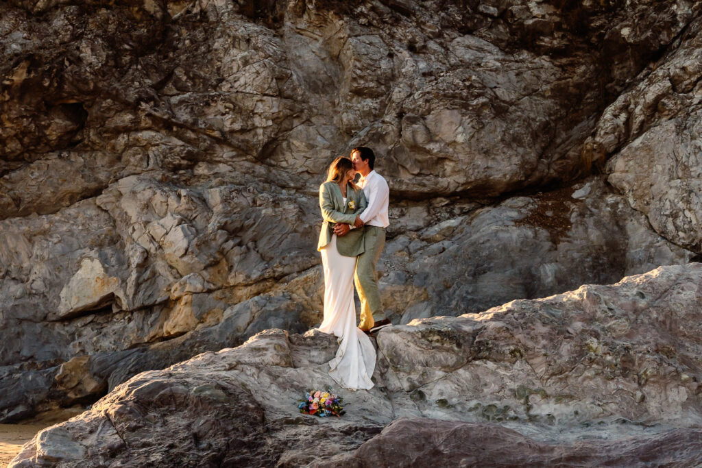 A bride and groom in wedding attire embrace on top of a rock during their coastal elopement