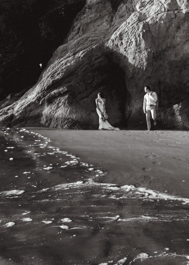 A black and white photo of a bride and groom casually leaning on a rock looking out at the ocean waves during their coastal elopement.