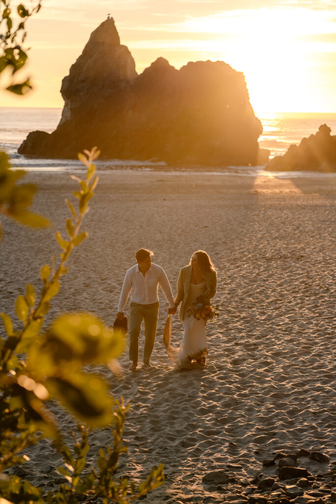 An image of a bride and groom exploring during their oregon coast elopement. They walk through a sandy beach with the sun flaring behind them. 