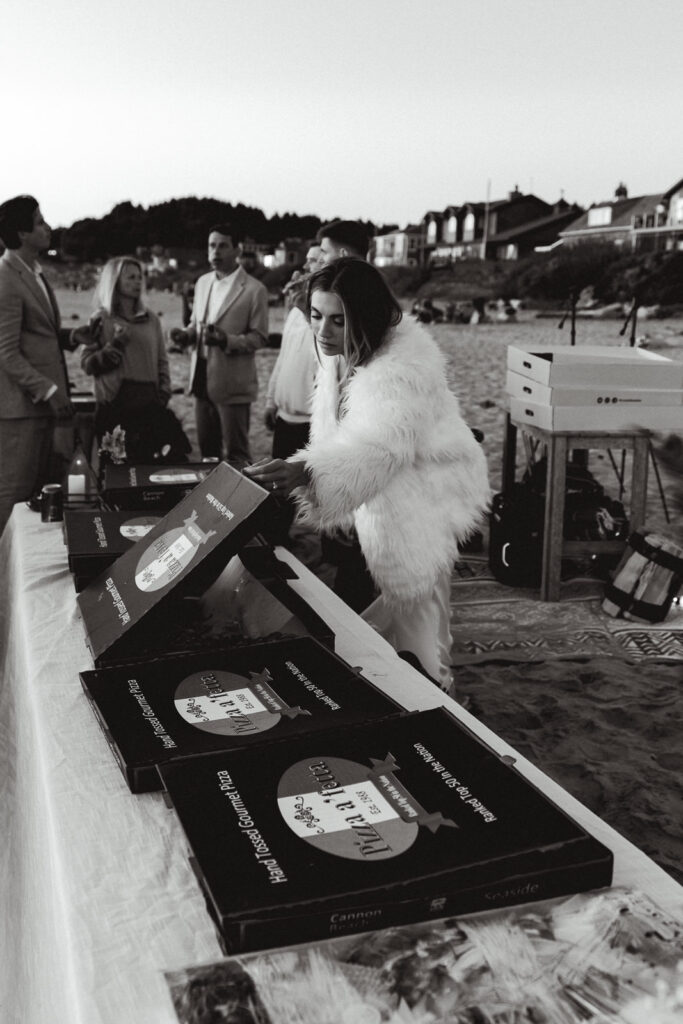 After her coastal elopement, a bride grabs pizza from the box during her bonfire beach reception.
