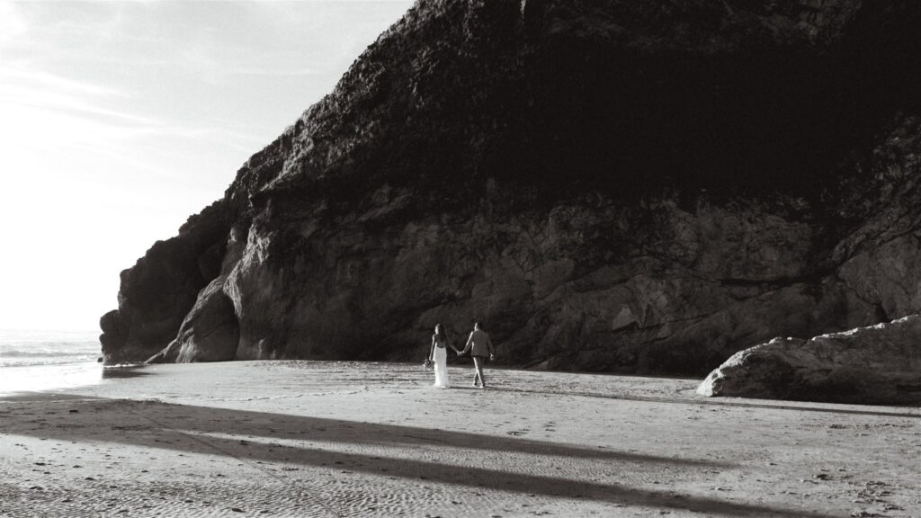During their coastal elopement, a bride and groom walk and explore a rocky beach, long shadows stretch out behind them.