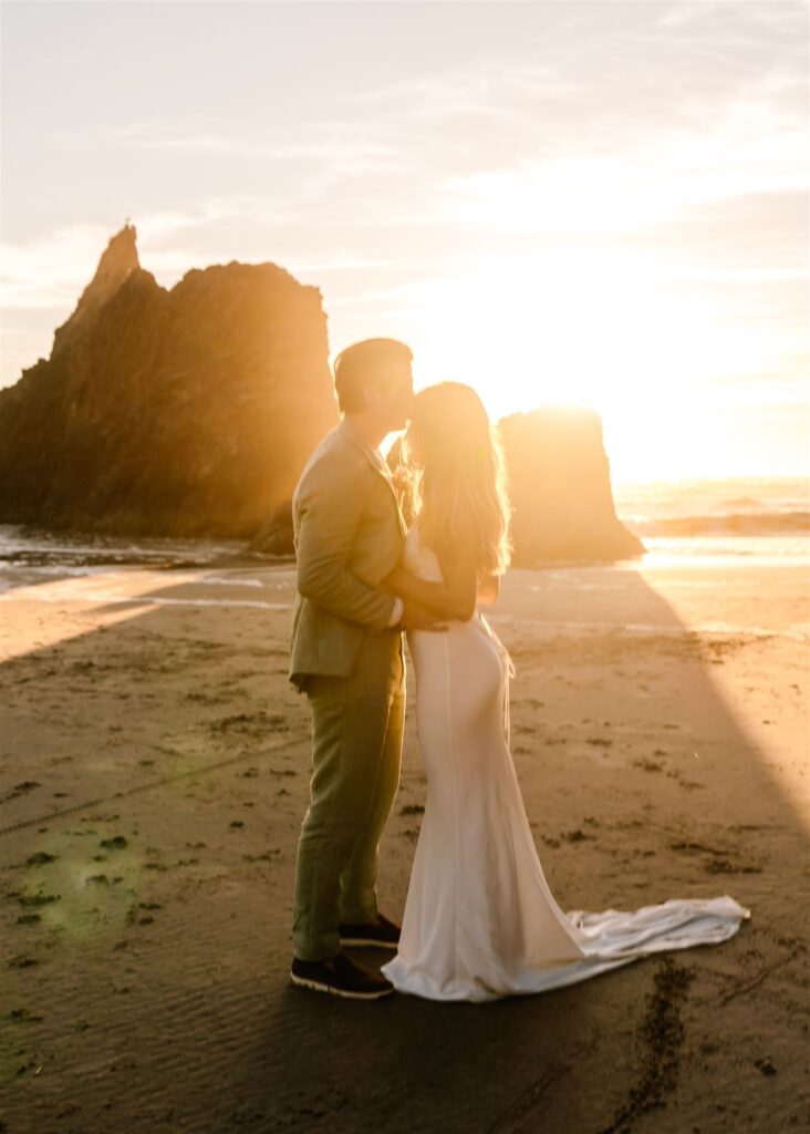 The sun flares behind a bride and groom on the beach as they embrace during their coastal elopement