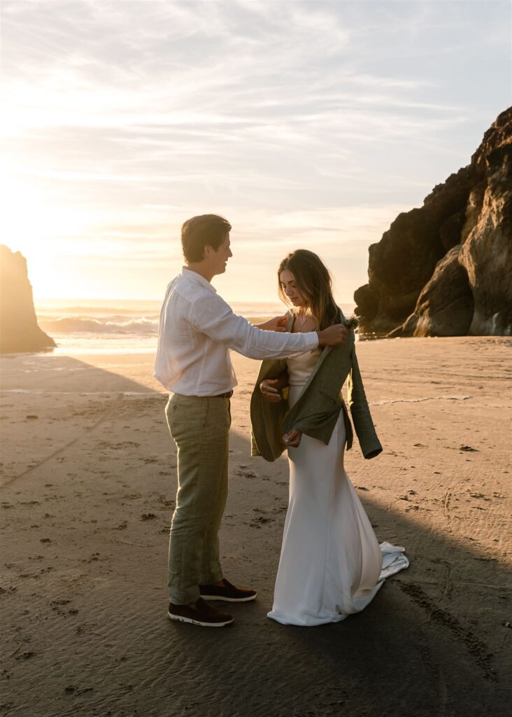 during their coastal elopement, a groom lends his bride his jacket. The sun flares behind them as they exchange loving looks. 