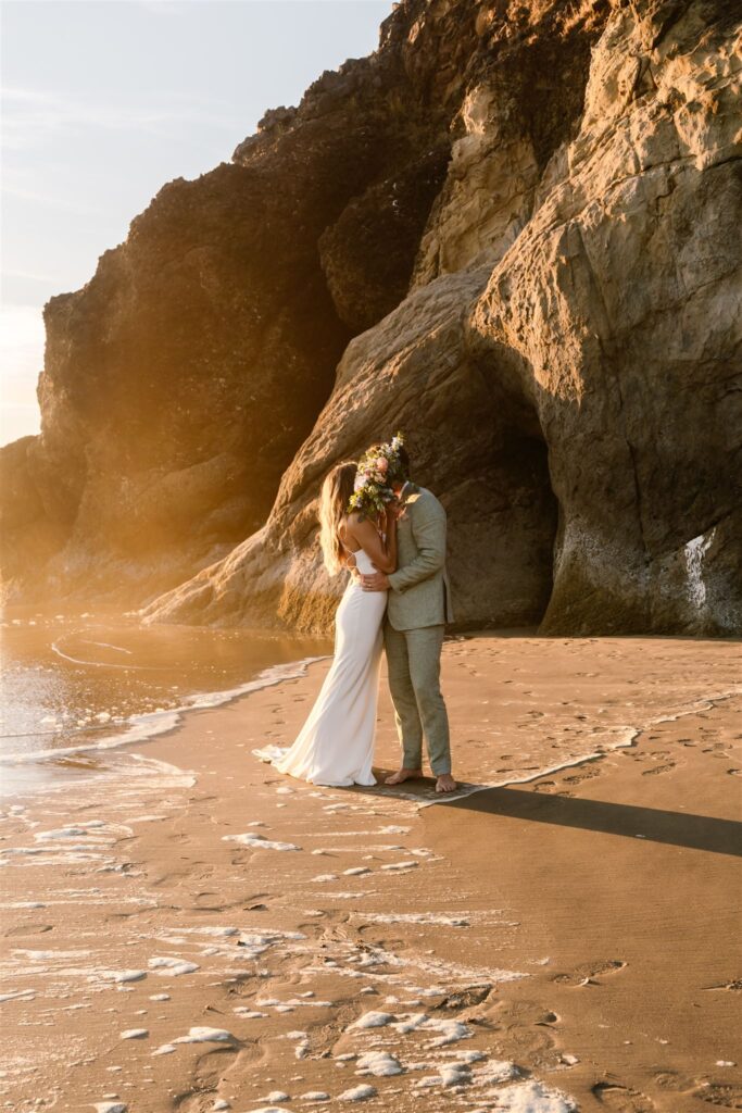 during their coastal elopement, a bride and groom kiss in front of a large sea stack. The bride holds her colorful bouquet over their faces.