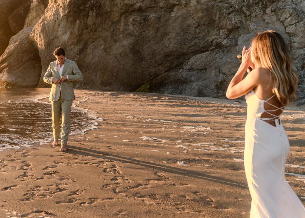 During their coastal elopement, a bride films her groom on a vintage super 8 camera as he adjusts his coat