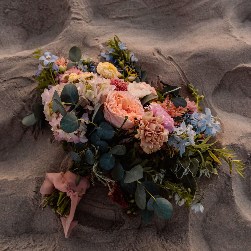 The bouquet of bright pinks, blues, and earthy greens lays in the sand during the celebration of their coastal elopement. 
