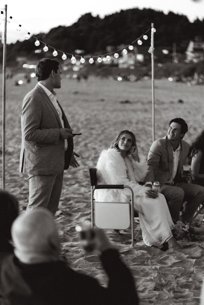 during the bonfire beach reception of this coastal elopement, a groomsman offers up a toast. the bride and groom smile as he speaks.