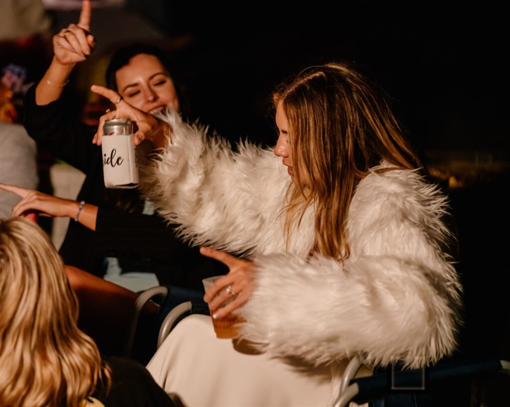 to celebrate her coastal elopement, a bride dances in her chair during her bonfire beach reception. She wears a furry coat and holds a beer in her hand as she vibes to the music.