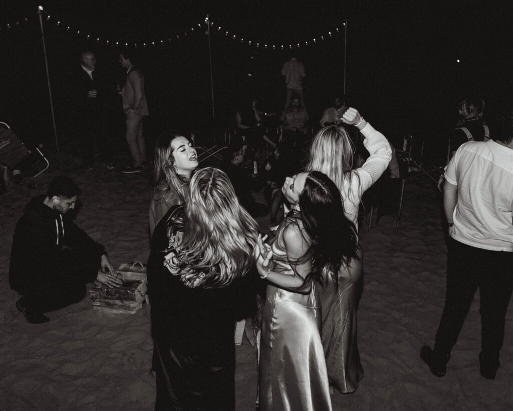 guests dance to the music at a bonfire beach reception to celebrate the bride and grooms coastal elopement. They wear jackets over their silk dresses and dance barefoot in the sand. 