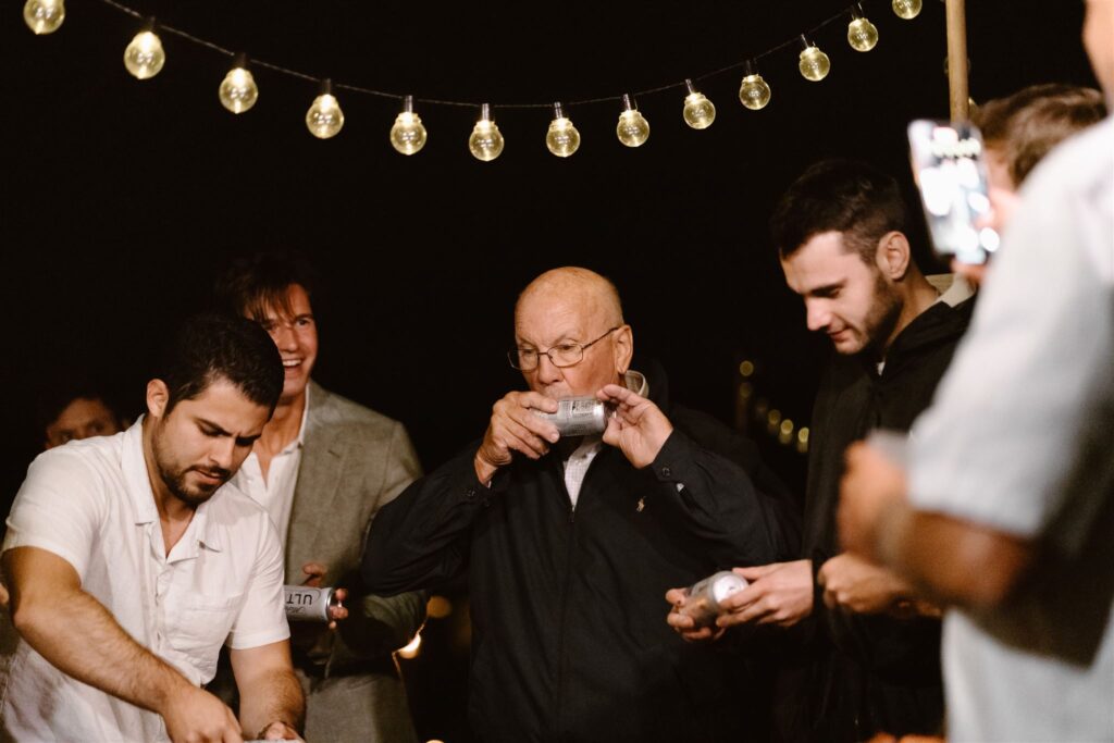 During the bonfire beach reception, guests shotgun beers to celebrate a coastal elopement. The brides grandfather stands center.