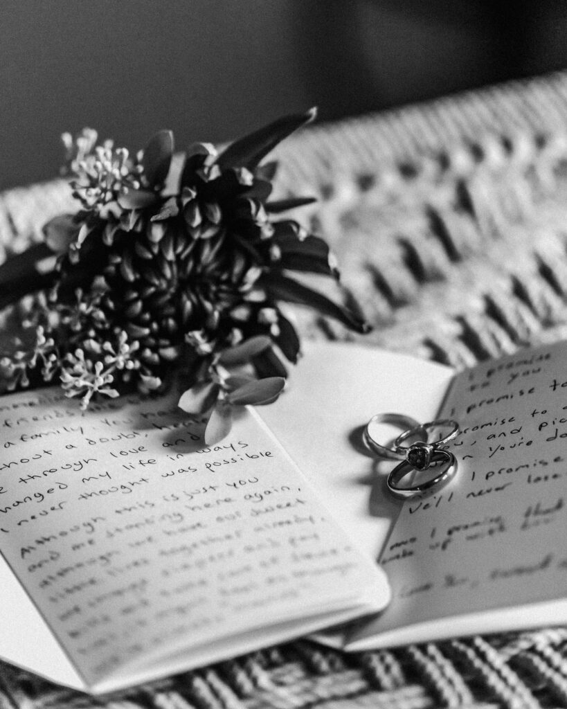 A black and white image of the details of this couples surfer wedding. Their details include their vow books, rings, and the grooms boutonnière. 