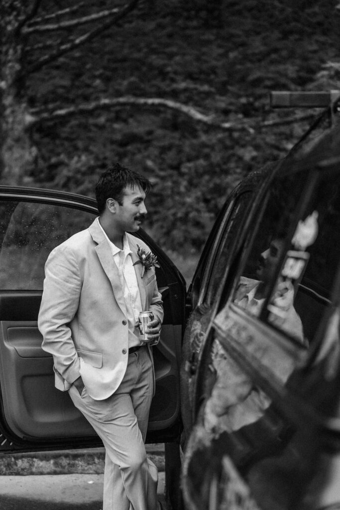 After their surfer wedding, a groom stands at the door of their car, warming up and enjoying a celebratory beer