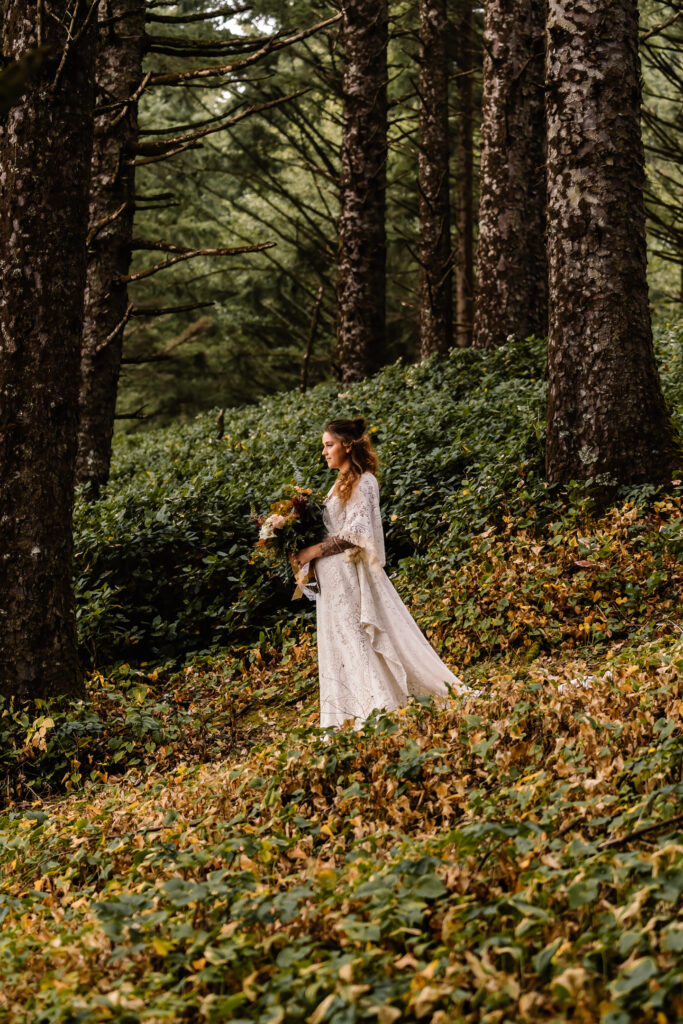 A bride walks through a coastal forest in her dress, holding her flowers as she prepares for her surfer wedding.