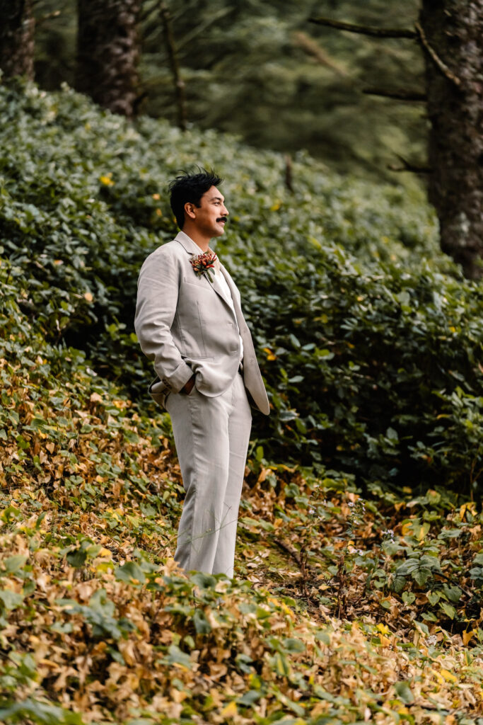 A groom stands in a coastal forest while wearing his suit for his surfer elopement