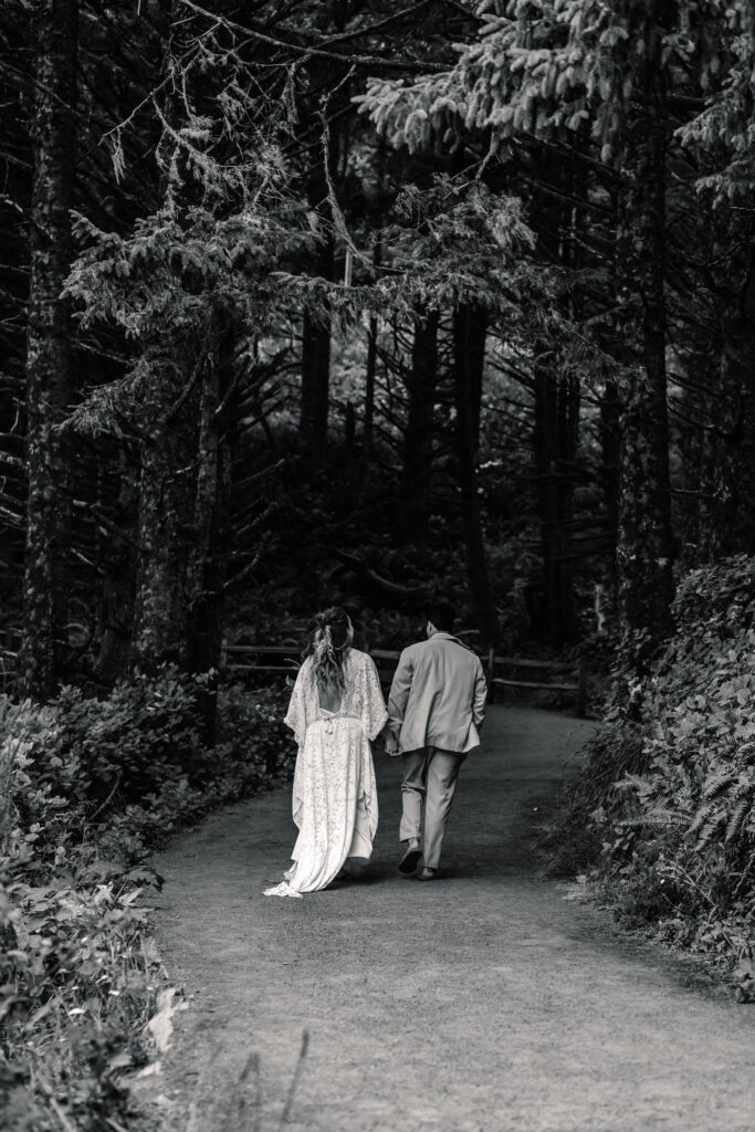 A black and white image of a bride and groom hiking through a coastal forest before their surfer wedding. The image is black and white, bringing highlight the shadows of the forest, drawing focus to the couple on their journey.