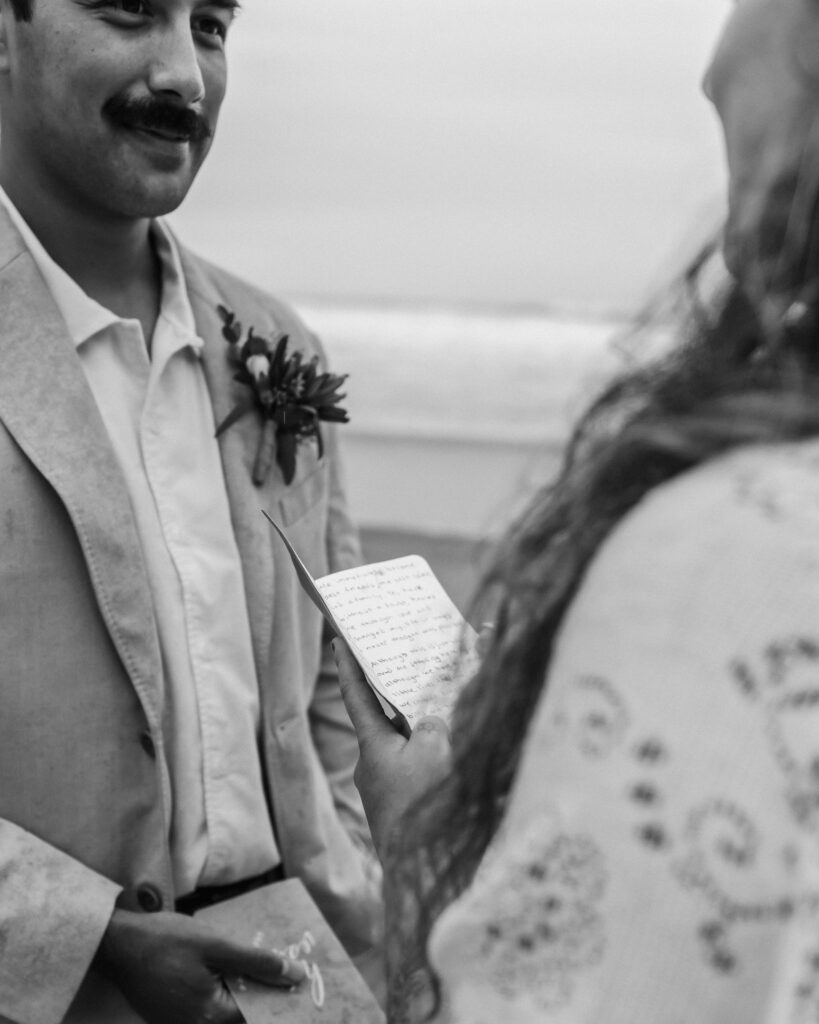A black and white image of a story telling shot of a brides vows. She reads them to her groom on a moody beach during their surfer wedding.